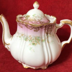 Peut inclure: Théière en porcelaine blanche avec un motif floral rose et doré. La théière a un bec verseur incurvé, une anse et un couvercle avec un bouton doré. Le corps de la théière est nervuré et les bords sont dorés.