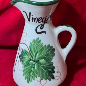 Könnte beinhalten: Weißer Essigkrug aus Keramik mit grünem Rand und Griff. Das Wort "Vinegar" ist in schwarzer Schrift geschrieben. Ein großes grünes Weinblatt mit braunen Ranken verziert die Vorderseite. Der Krug hat einen Ausguss.