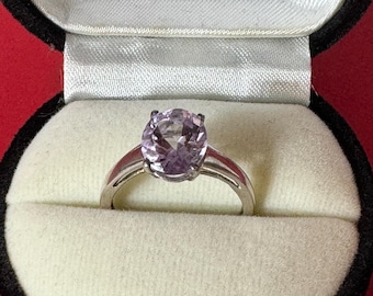 May include: A silver ring featuring a large, round, light purple gemstone. The ring is presented in a velvet-lined box with a black exterior and a red base. The gemstone is the central feature.