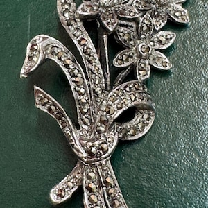May include: A silver-toned marcasite brooch in the shape of a floral bouquet. The brooch features three flowers and a ribbon, all encrusted with small, sparkling marcasite stones. The design is detailed and elegant, suitable for adding a touch of vintage charm.