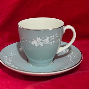 Op de afbeelding: Een lichtblauwe theekop en schotel set met een wit bloemen- en esdoornbladpatroon. De kop heeft een witte binnenkant en een zilveren rand, met een bijpassende schotel met zilveren rand. Het ontwerp bevat delicate witte takjes en bladeren.