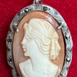 Op de afbeelding: Ovale hanger met een camee van een vrouwenprofiel in crème tegen een perzikkleurige achtergrond. De camee is gezet in een zilverkleurig montuur met decoratieve details en kleine, donkere accenten. De hanger hangt aan een zilveren lus, tegen een rode achtergrond.