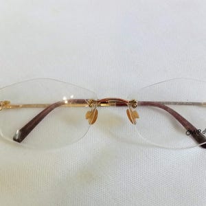 Puede incluir: Gafas sin montura con montura de metal dorado y patillas marrones. Las lentes son transparentes y tienen una forma geom&eacute;trica sutil. El texto "OWP design" est&aacute; impreso en la lente inferior derecha.