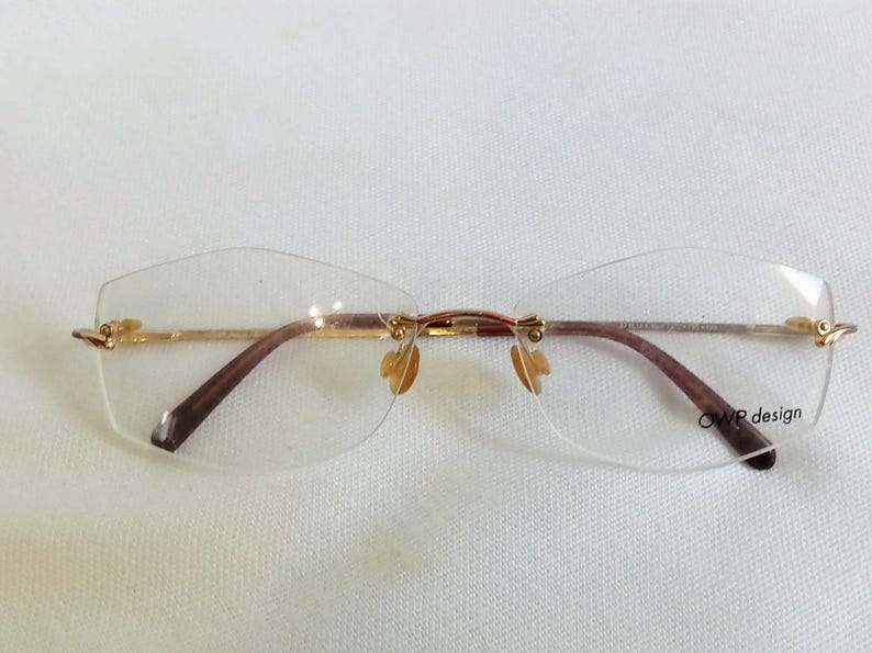 Puede incluir: Gafas sin montura con montura de metal dorado y patillas marrones. Las lentes son transparentes y tienen una forma geom&eacute;trica sutil. El texto "OWP design" est&aacute; impreso en la lente inferior derecha.