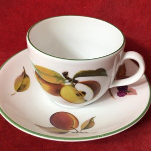 Puede incluir: Juego de taza y platillo blancos con ribete verde. La taza y el platillo están decorados con ilustraciones de manzanas, peras y ciruelas. La taza tiene un asa y se apoya en el platillo a juego. El juego está sobre un fondo rojo.