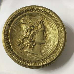 Könnte beinhalten: Eine goldfarbene Metallmedaille mit einem Reliefbild eines Kopfes einer Person, die einen Kranz trägt. Die Medaille hat einen dekorativen Rand.
