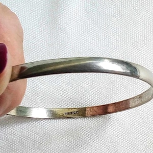 Peut inclure: Un bracelet jonc de couleur argentée. Le bracelet est rond et lisse, avec une surface légèrement incurvée. Le mot "MEXICO" est estampillé à l'intérieur du bracelet. Le bracelet est tenu par une main avec du vernis à ongles violet foncé.