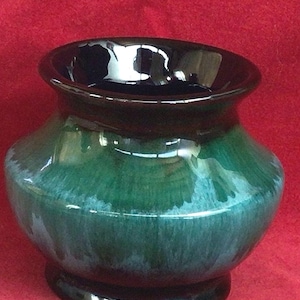 Puede incluir: Un jarrón de cerámica brillante en verde oscuro y negro. El jarrón tiene un cuerpo ancho y redondeado y un borde acampanado. La base tiene una forma circular oscura. El jarrón mide unos 15 cm de alto.