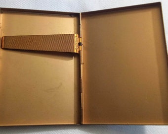 May include: A gold-colored metal cigarette case with a hinged lid and a gold-colored metal clasp. The case is open and the inside is empty.