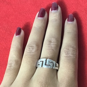 Könnte beinhalten: Ein silberner Ring mit einem griechischen Schlüsselmuster. Der Ring wird an einem Finger mit dunkelrotem Nagellack getragen.