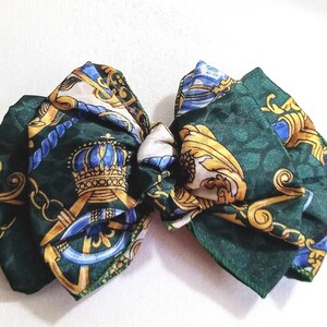 May include: A dark green bow with a repeating pattern of gold, blue, and white nautical and crown designs. The bow is made of a silky material and is tied in the centre. The bow is on a white background.