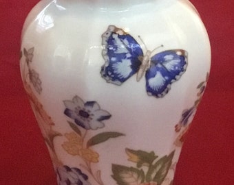 Puede incluir: Un jarrón de porcelana blanca con un borde festoneado y un diseño floral. El jarrón presenta una mariposa azul con detalles dorados y una rosa rosa con hojas verdes.
