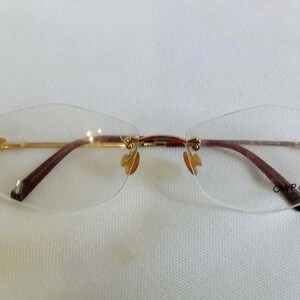 Puede incluir: Gafas sin montura con puente y varillas doradas y patillas marrones. Las lentes son transparentes y tienen un dise&ntilde;o geom&eacute;trico sutil. Las almohadillas nasales son de color dorado claro. El texto "OWP design" es visible.