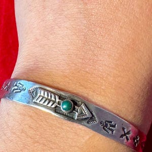 May include: Silver cuff bracelet with etched Native American-inspired designs. The bracelet features an arrow and a small, round, green stone. The cuff is worn on a wrist against a red background.