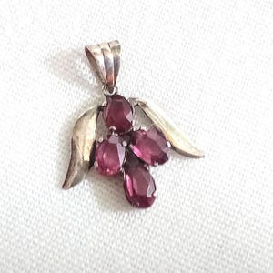 May include: A silver pendant featuring a cluster of oval, deep red gemstones. The pendant has a stylized leaf design on either side of the gemstones and a loop at the top for a chain. The gemstones are a rich, dark red colour.