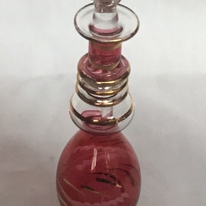 Può includere: Una bottiglia di vetro rosa con accenti dorati e un tappo di vetro trasparente. La bottiglia ha un design a spirale e poggia su una base rosa.