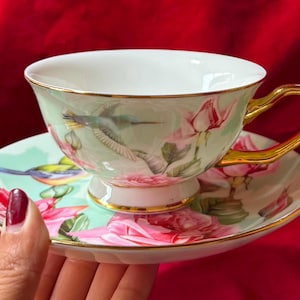 Op de afbeelding: Een delicate theekop en schotel set. De kop en schotel zijn versierd met roze rozen, kolibries en gouden randen. De kop heeft een gouden oor. De achtergrond is rood.