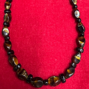 Op de afbeelding: Een ketting met ovale tijgeroog kralen en kleine zwarte kralen. De tijgeroog kralen hebben bruine, gouden en zwarte tinten. De ketting is afgebeeld tegen een rode achtergrond.
