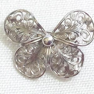 Peut inclure: Broche papillon argentée avec des détails de filigrane complexes. La broche présente une perle centrale et quatre ailes symétriques, chacune avec des motifs délicats et tourbillonnants. Le métal a une finition polie.