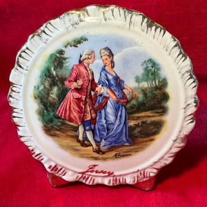 May include: A decorative ceramic plate with a scalloped edge and gold trim. The plate features a painted scene of two figures in period clothing, with the word "Jersey" written at the bottom.
