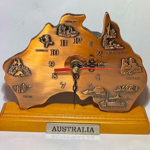Pode incluir: Um relógio cor de cobre em forma do continente australiano, com nomes de cidades e marcos gravados. Possui uma base de madeira e uma placa de metal com a inscrição "AUSTRALIA". Os ponteiros do relógio são vermelhos.