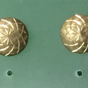 May include: A pair of gold-toned earrings with a geometric design. The earrings are round and have a textured surface.