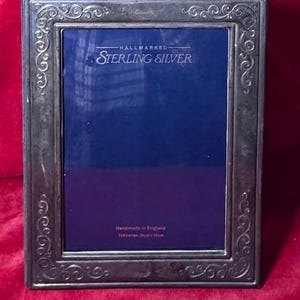 May include: A rectangular sterling silver picture frame with ornate scrollwork around the edges. The frame has a dark blue interior and the text "HALLMARKED STERLING SILVER" is visible. The frame is 7x5 inches (18cm x 13cm).
