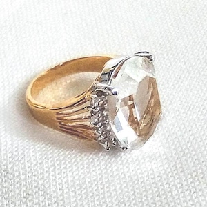 May include: A gold ring featuring a large, clear, faceted gemstone. The oval-shaped gemstone is set in a silver and gold band. The ring is shown on a white, textured surface.