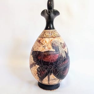 May include: An antique, egg-shaped ceramic vase with a black handle and base. The vase features a painted design of a figure in red and black, with the word "SHINA" visible. The background is beige.