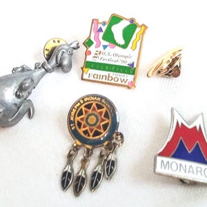 May include: A selection of vintage enamel pins. Includes a silver frog pin, a gold pin, a pin with the text "U.S. Olympic Festival '90", a pin with the text "St. Joseph's Indian School", and a pin with the text "MONARCH".