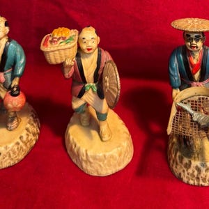Puede incluir: Tres figuras japonesas vintage que representan figuras con vestimenta tradicional. Las figuras están pintadas con colores vibrantes y se apoyan sobre bases texturizadas. Una figura lleva una linterna, otra una cesta de frutas y la tercera una red de pesca.
