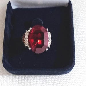 May include: A striking ring featuring a large, oval-cut, red gemstone, set in a silver band, presented in a dark blue velvet box. The gemstone is the main feature, reflecting light. A statement ring.