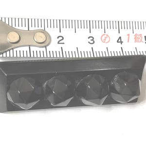 May include: A black rectangular object with four faceted, gem-like protrusions. The object is approximately 1.75 inches long, as measured by a metal ruler in the image. The object is likely a decorative element or hardware.