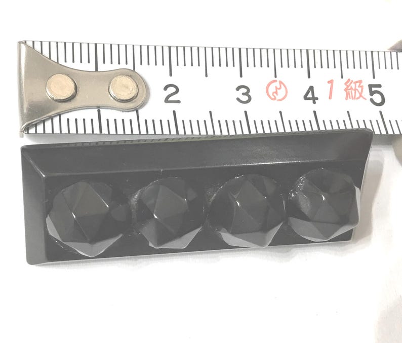 May include: A black rectangular object with four faceted, gem-like protrusions. The object is approximately 1.75 inches long, as measured by a metal ruler in the image. The object is likely a decorative element or hardware.