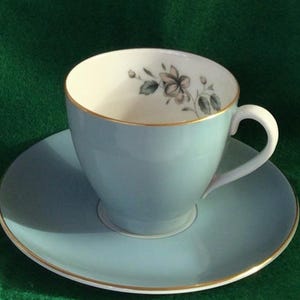 Può includere: Un set da tè composto da una tazza e un piattino di colore azzurro chiaro con un motivo floreale. La tazza ha un bordo dorato e il piattino ha una fascia dorata intorno al bordo.