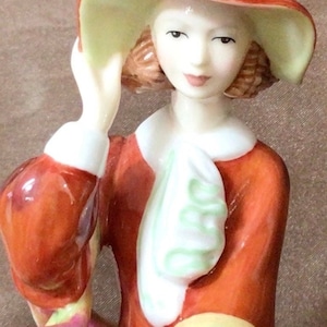 May include: A porcelain figurine of a woman wearing a red and white dress with a green scarf. She is wearing a red hat with a blue ribbon and has her hand on her hat.