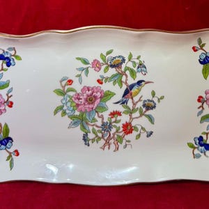 Puede incluir: Una bandeja de servicio rectangular de cerámica con bordes festoneados y adornos dorados en las asas. La superficie blanca presenta un arreglo floral central con un pequeño pájaro azul y amarillo, flanqueado por dos motivos florales.