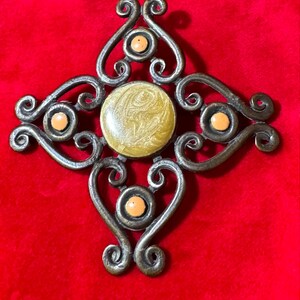 May include: Ornate, antique-style metal pendant with a central yellow-gold circular design and four smaller orange-yellow circles. The pendant has a symmetrical, cross-like shape with swirling, heart-shaped details. The background is a solid red.