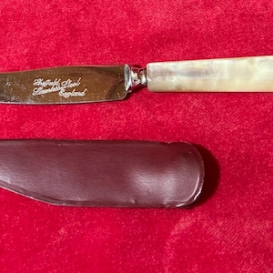 Puede incluir: Un cuchillo de mantequilla con una hoja de acero inoxidable y un mango nacarado. La hoja está grabada con "Sheffield Steel Stainless England". Se incluye una funda de cuero marrón.