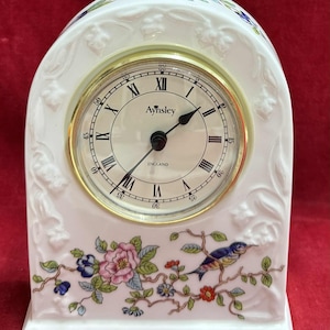 Puede incluir: Un reloj de chimenea de cerámica blanca con esfera dorada. El reloj presenta números romanos y la marca "Aynsley". Motivos florales y de pájaros decoran la base del reloj. El reloj mide aproximadamente 15 cm de altura.