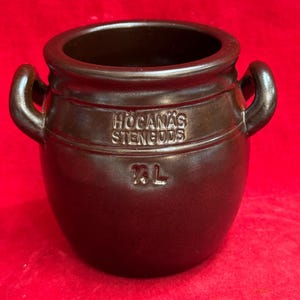 May include: A brown stoneware pot with two curved handles and the text "HOGANAS STENGODS" and "2L" embossed on the front. The pot has a wide opening and a rounded body. The pot is likely used for cooking or storage.