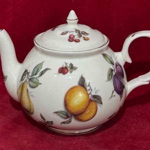 Könnte beinhalten: Eine weiße Teekanne aus Porzellan mit einer farbenfrohen botanischen Illustration verschiedener Früchte wie Birnen, Pflaumen und Kirschen. Die Kanne hat einen passenden Deckel mit spitzem Knauf, einen geschwungenen Henkel und einen Ausguss.
