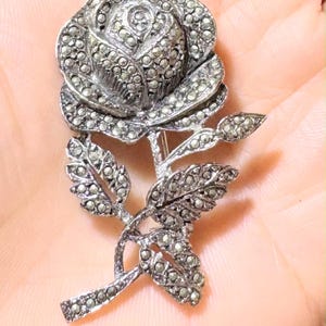 Op de afbeelding: Zilverkleurige broche in de vorm van een roos met gedetailleerde bloemblaadjes en bladeren. De broche is versierd met kleine, sprankelende steentjes, wat een textuur en sierlijk uiterlijk geeft. Het ontwerp is een klassiek bloemmotief.