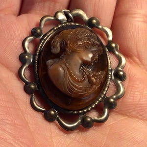 May include: A brown cameo pendant with a detailed profile of a woman in a dress with a pearl necklace. The cameo is set in a silver-toned ornate frame with a scalloped edge.