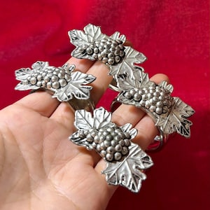 Könnte beinhalten: Set mit vier silberfarbenen Serviettenringen. Jeder Ring hat ein detailliertes Trauben- und Blattdesign. Die Ringe sind aus Metall und eignen sich für formelle Anlässe.