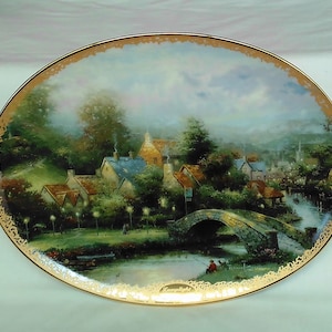 Peut inclure: Une assiette en céramique décorative avec une peinture représentant une scène de village pittoresque. L'assiette présente un pont au-dessus d'une rivière, des maisons, des arbres et une personne à la pêche. L'assiette a une bordure dorée et une inscription dorée qui dit "Thomas Kinkade".