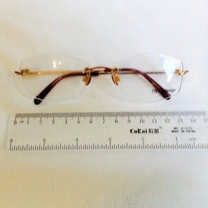 Puede incluir: Gafas sin montura con detalles met&aacute;licos dorados y patillas marrones. Las gafas est&aacute;n junto a una regla, con el texto "CoKai" visible. Las gafas miden aproximadamente 14 cm de ancho.
