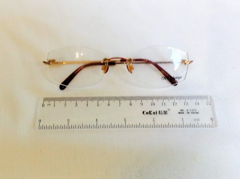 Puede incluir: Gafas sin montura con detalles met&aacute;licos dorados y patillas marrones. Las gafas est&aacute;n junto a una regla, con el texto "CoKai" visible. Las gafas miden aproximadamente 14 cm de ancho.