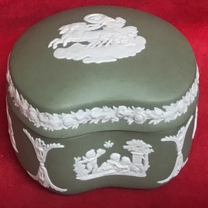 Può includere: Una scatola portaoggetti in ceramica verde con disegni in rilievo bianchi. Il coperchio presenta un carro trainato da un cavallo e il fondo presenta una scena con tre figure.