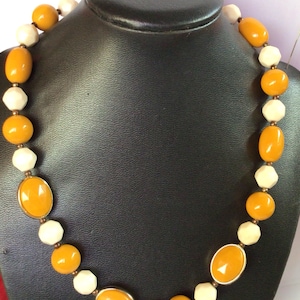 May include: A necklace made of alternating oval-shaped amber and white beads. The necklace is displayed on a black mannequin.